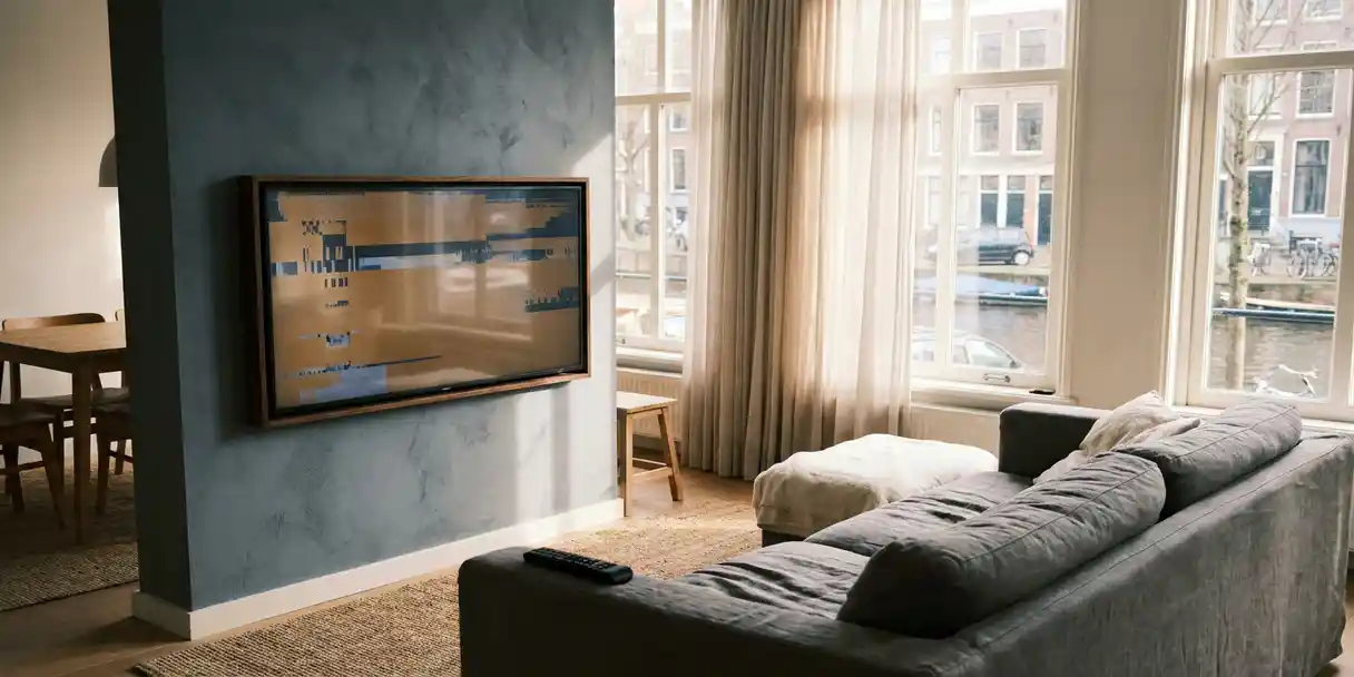 televisie-storing-woonkamer-amsterdam-interieur.jpg Flatscreen tv aan de muur van een Amsterdamse woonkamer met een bevroren reclamebeeld, afstandsbediening op een grijze linnen bank.