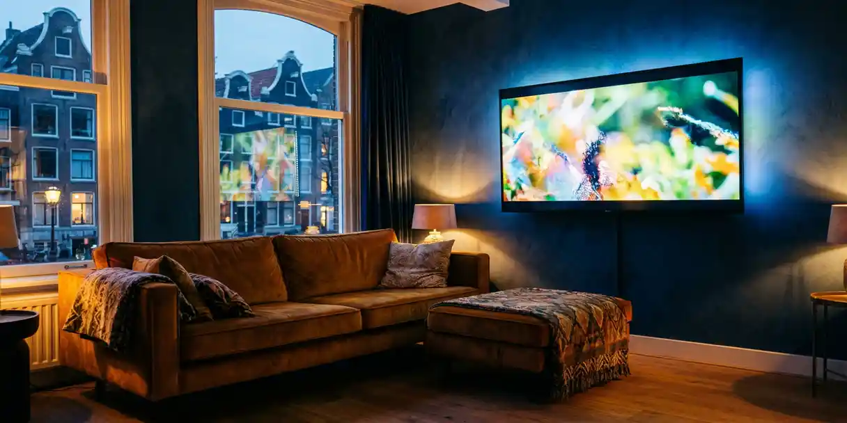 Flatscreen televisie met streaming content in een Amsterdamse woonkamer met kanaalhuisramen en uitzicht op daken in de schemering.